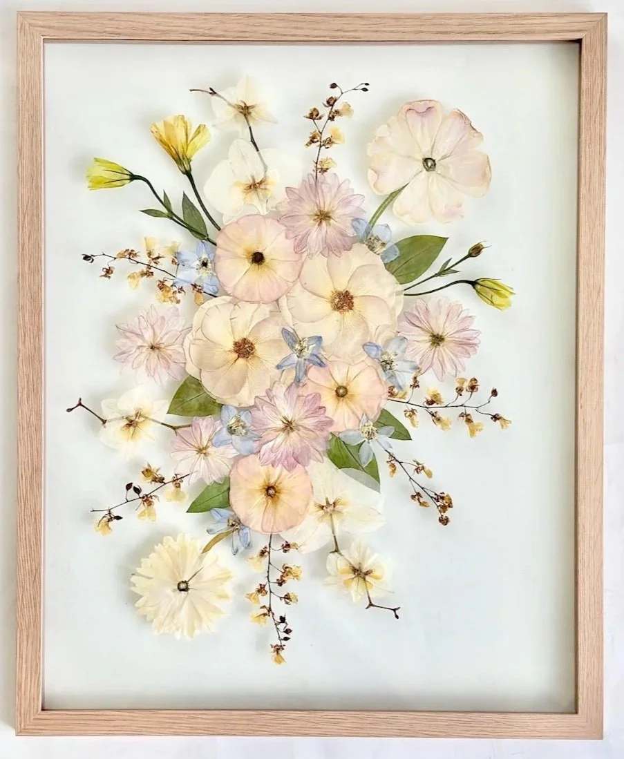 Pressed flower art with pastel-colored flowers, leaves, and small yellow and blue blossoms in a wooden frame.