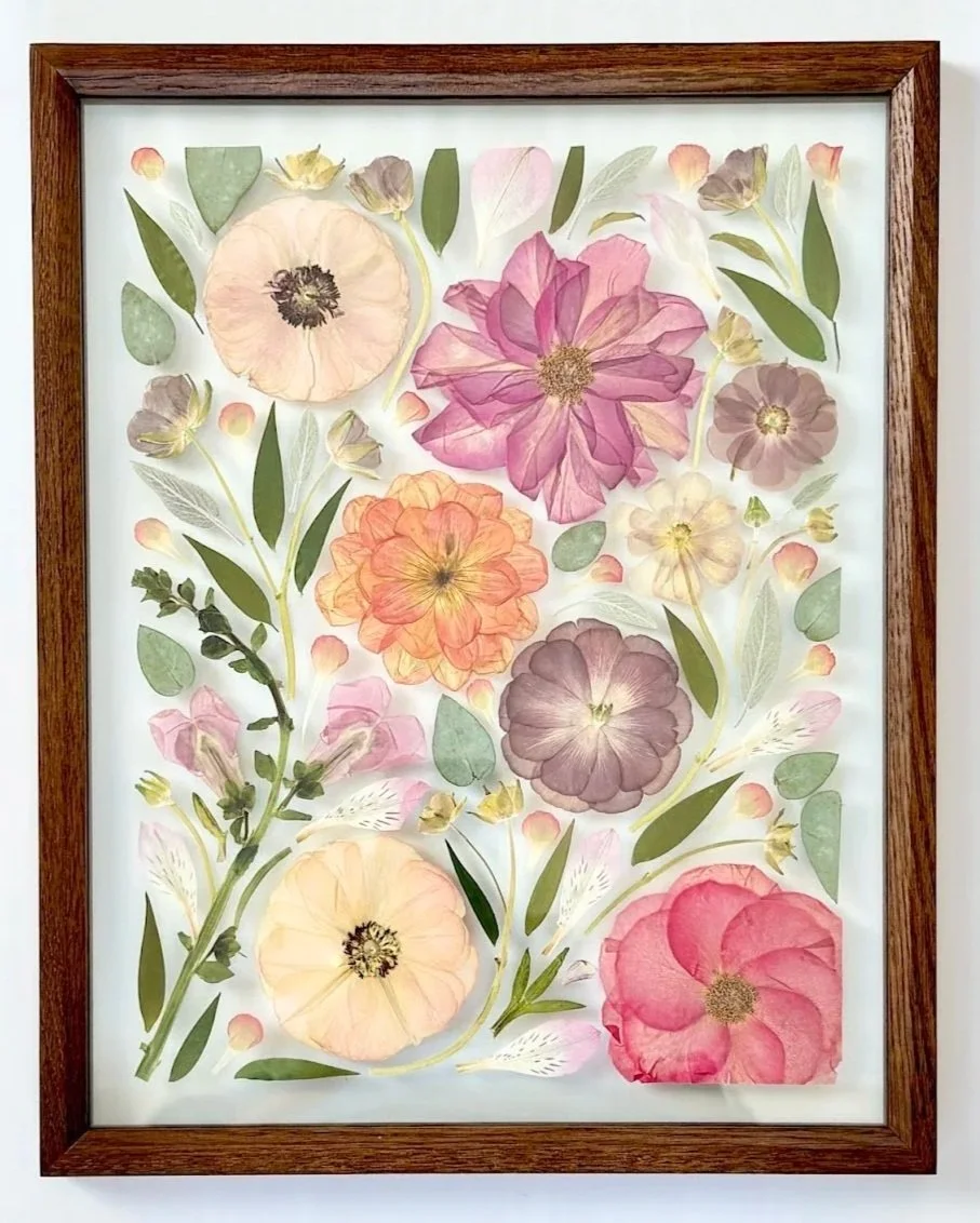 Framed artwork of pressed flowers with pink, purple, peach, and cream blossoms, green leaves, and small buds on a white background.