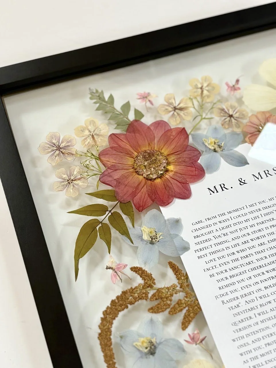 Framed wedding vow with pressed flowers including a large red and yellow flower, blue, pink, and beige flowers, and green leaves.