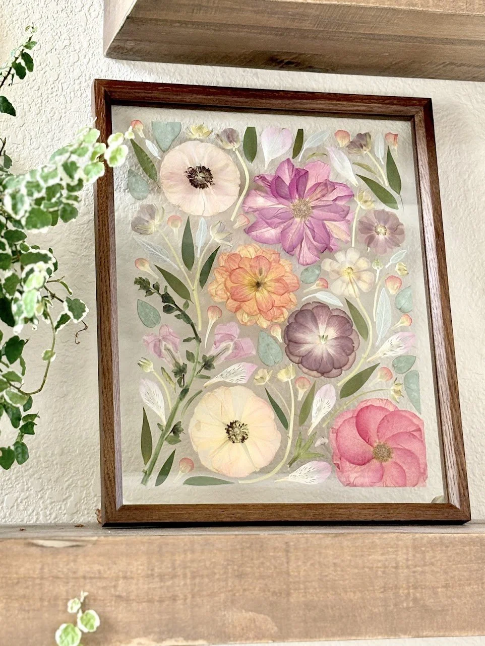 Framed artwork of various colorful, watercolor-style flowers and leaves on a white background, hanging on a textured wall.