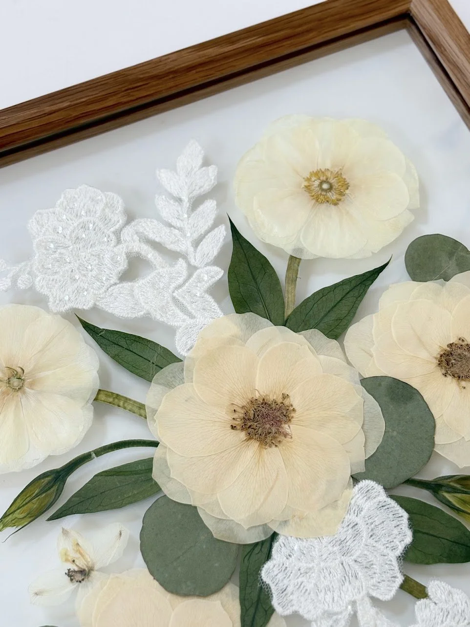 Pressed cream-colored flowers, green leaves, and white embroidered lace leaves arranged inside a wooden frame.