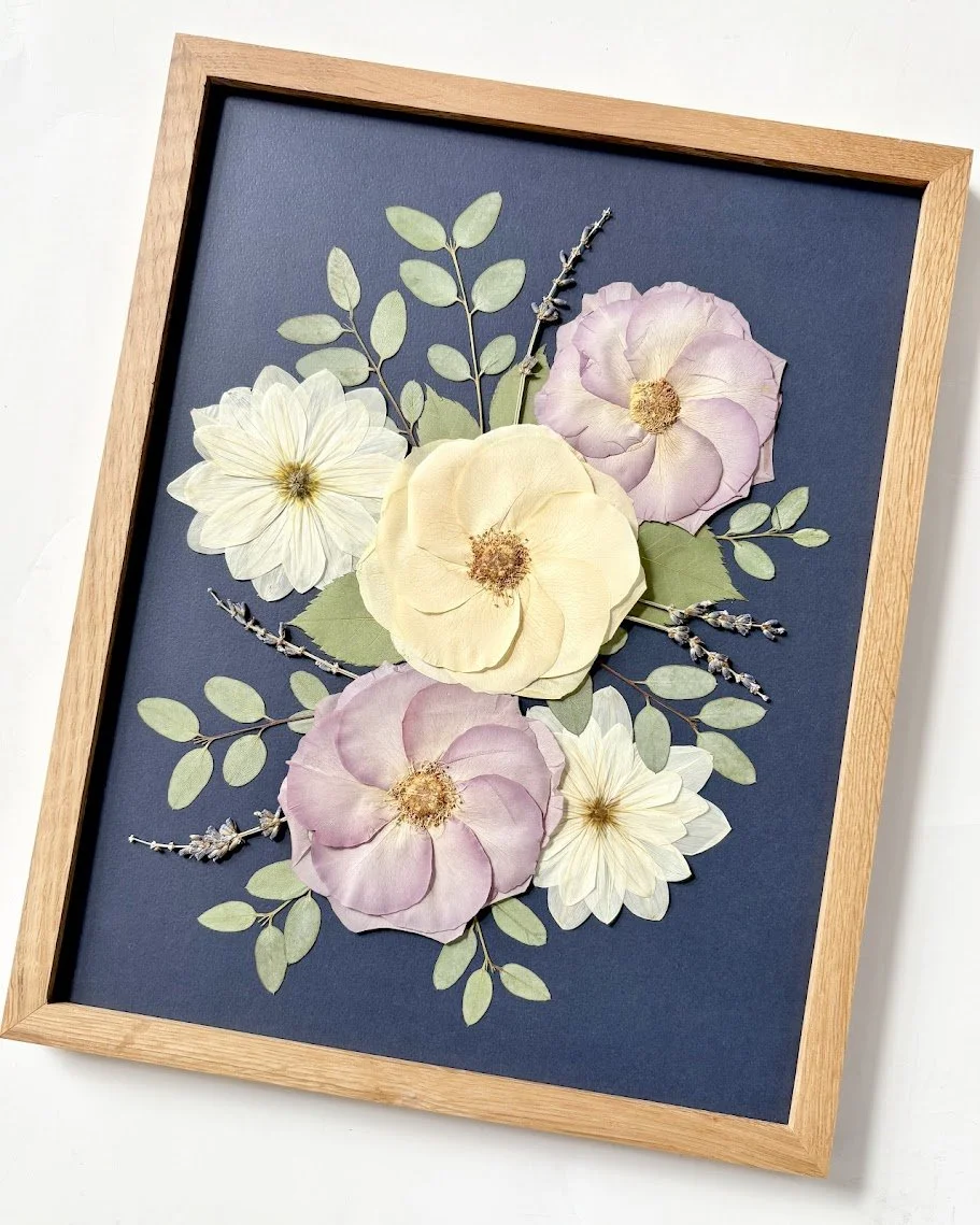 Pressed floral art framed in a wooden frame, featuring pink, yellow, and white flowers with green leaves and sprigs, set against a dark blue background.