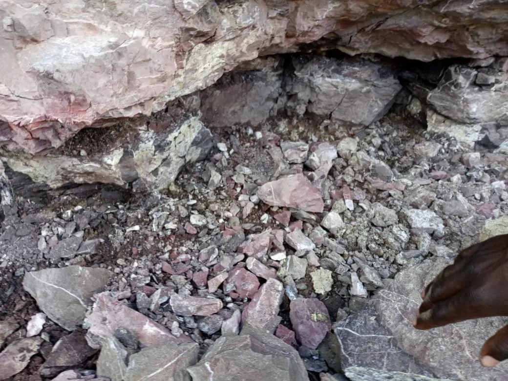 Close-up of a dirt and rock area, with a crevice or small excavation site in a rocky terrain. Some rocks and dirt are visible, with parts of a person's finger in the bottom right corner.