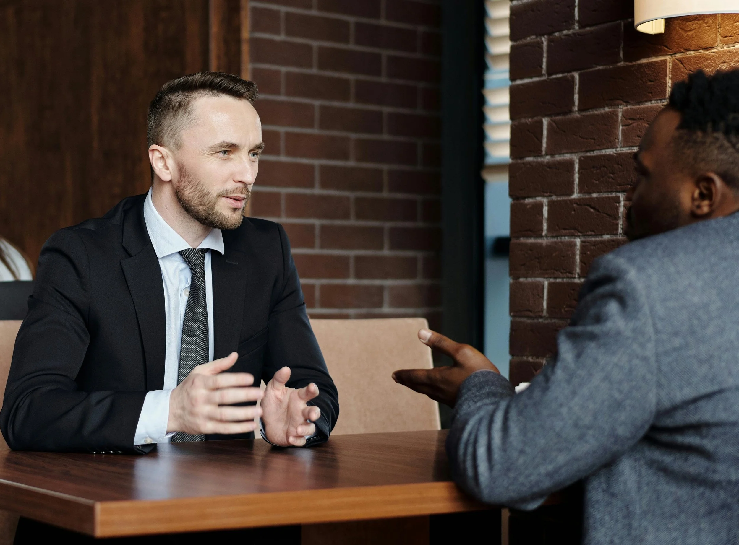 Two men engaging in a conversation at a table in a brick-walled room.