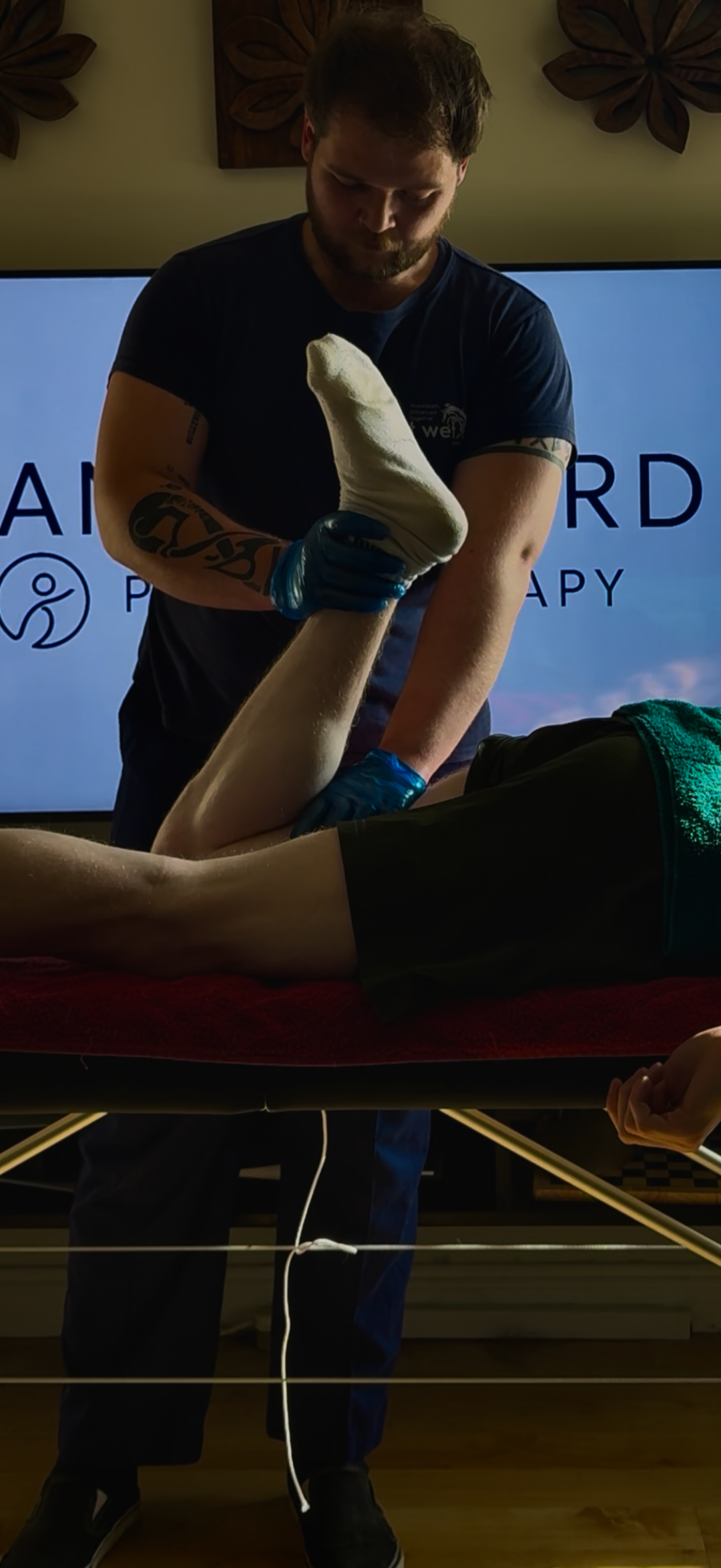 A person performing physical therapy or stretching exercises on a leg while lying on a treatment table, assisted by another individual, with a screen in the background displaying the word 'therapy'.