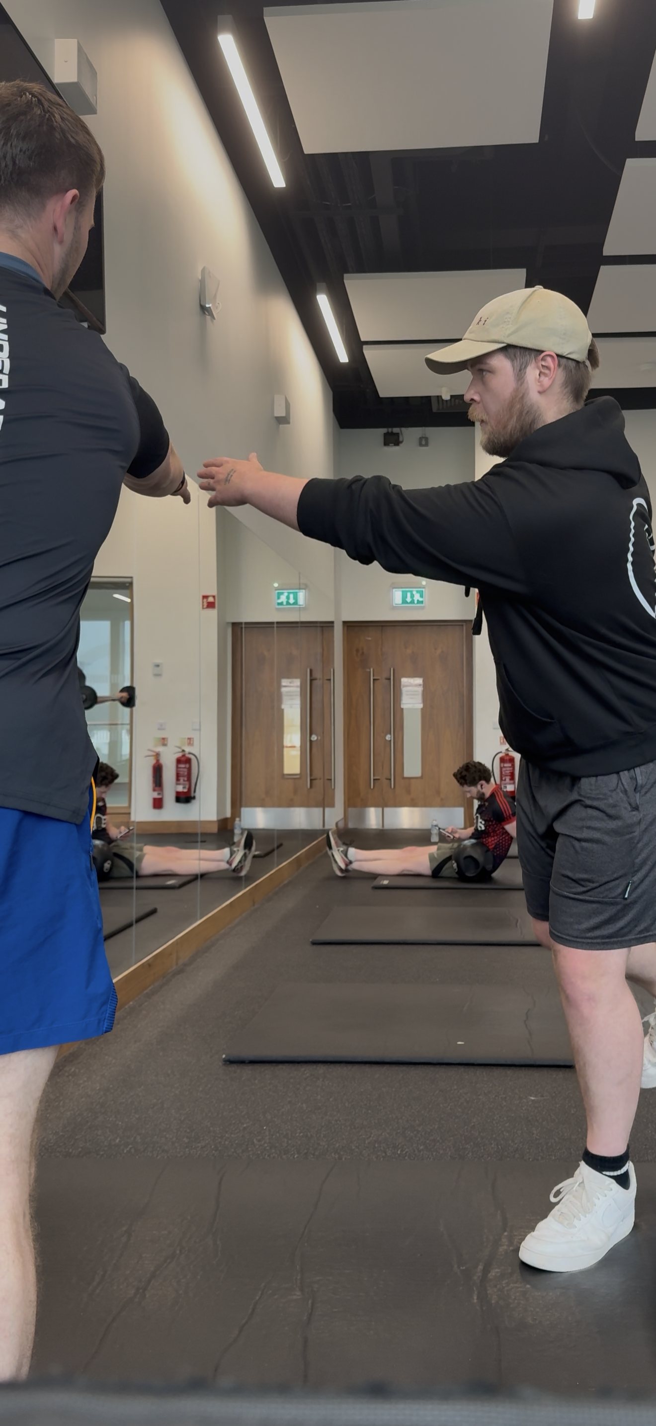 Two men are in a gym, with one man explaining or demonstrating something to the other, who is sitting on the floor stretching. The gym has a mirrored wall, exercise mats, and two other people in the background, one sitting with a water bottle and the other lying on an exercise mat.