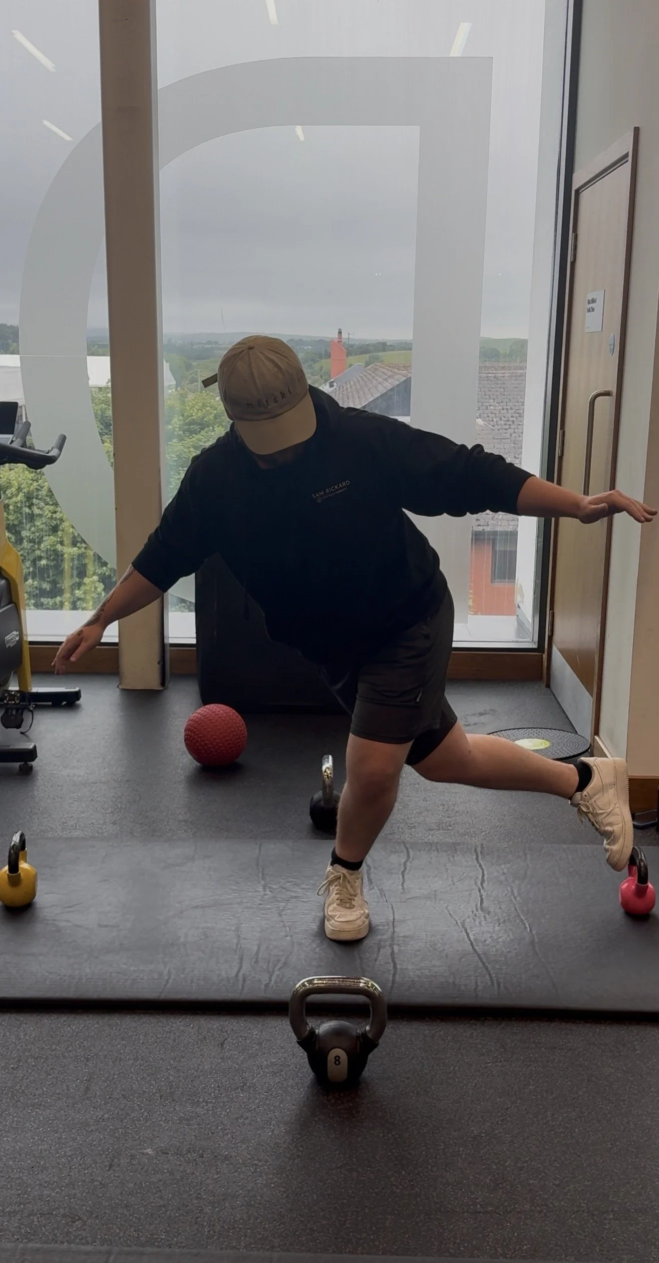 Person in gym attire performing a balance exercise on one leg, with arms extended, near exercise equipment and a large window showing an overcast sky and rooftops outside.