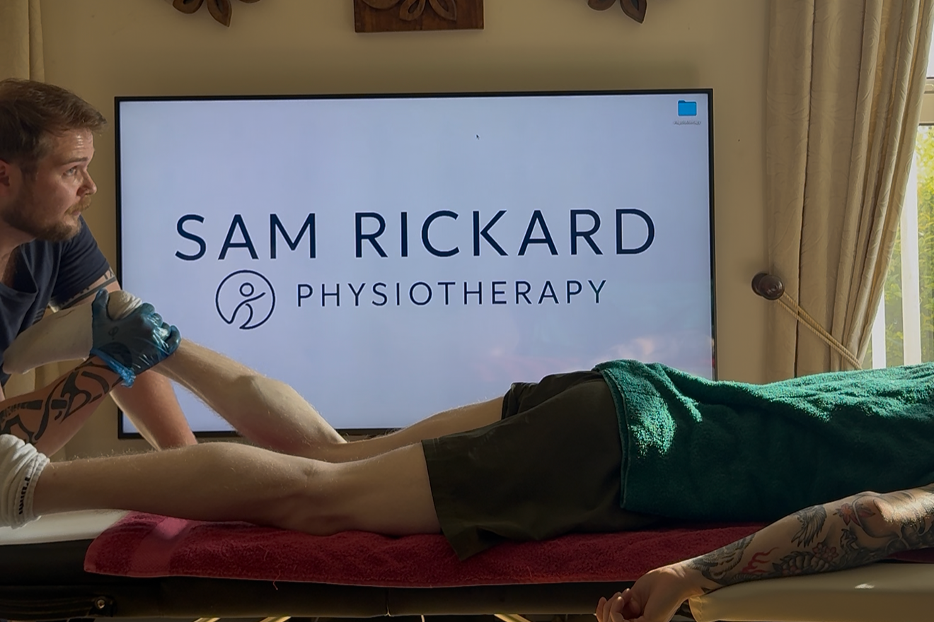 A physiotherapist performs a physical therapy session on a patient lying face down on a treatment table. The physiotherapist holds the patient's leg while another practitioner supports the patient. A large screen in the background displays the logo and name 'Sam Rickard Physiotherapy'.
