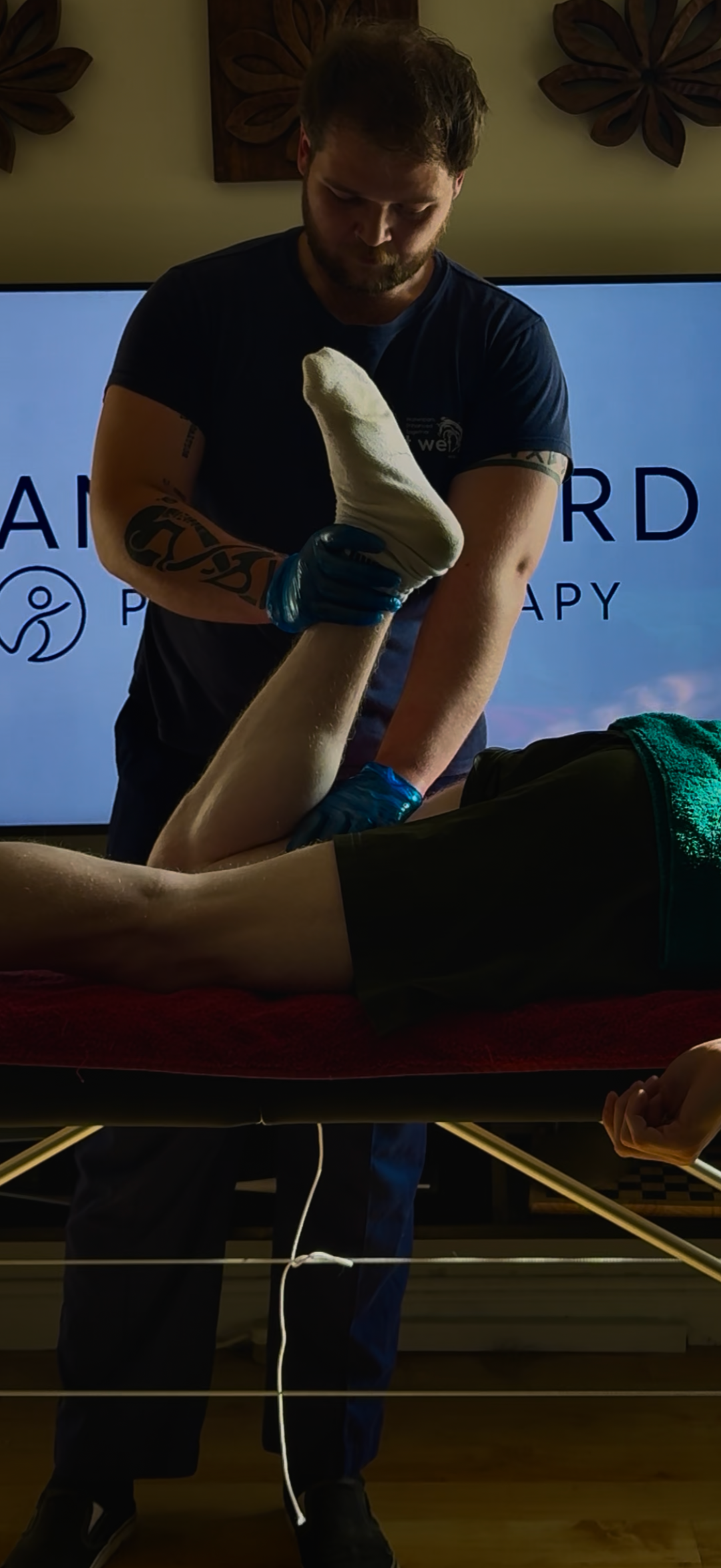 A man receiving physical therapy on his leg, lying on an examination table, with a therapist holding his leg and ankle. The background displays a screen with therapy-related text and a logo.