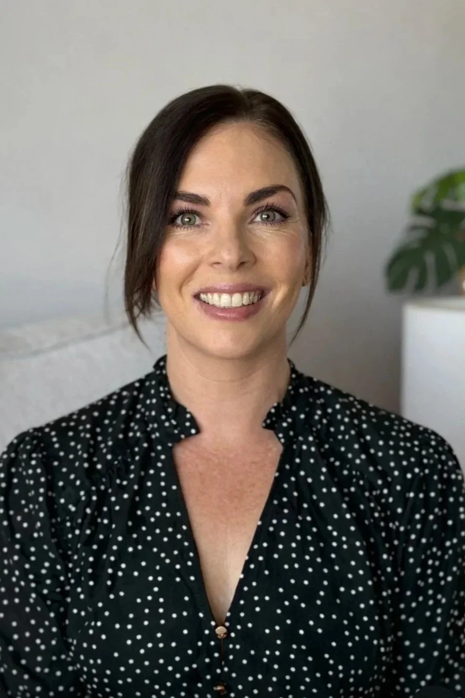 Portrait of a woman with dark brown hair, light eyes, and a friendly smile, wearing a black blouse with white polka dots, in a well-lit room with a neutral background and a plant on the right.
