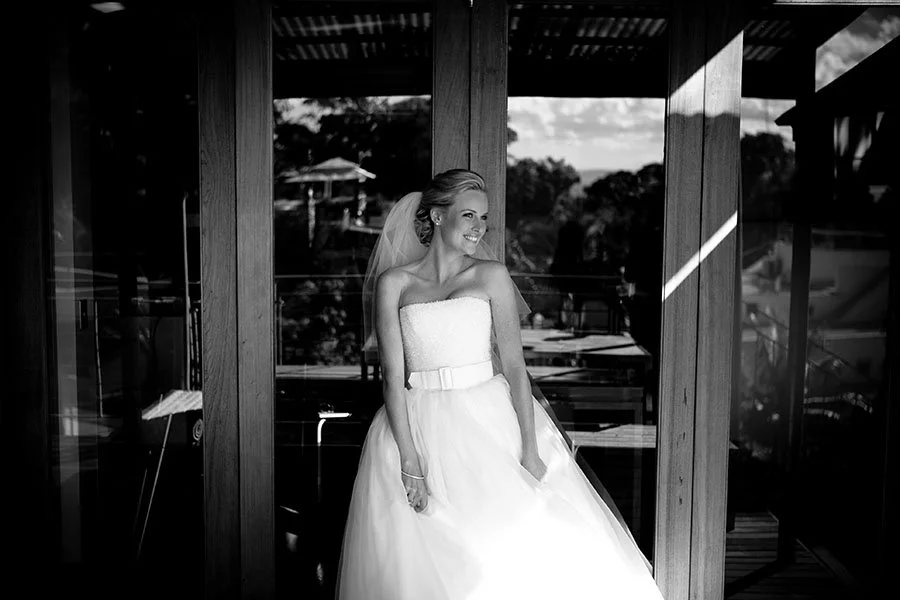 A bride in a strapless wedding dress with a belt, smiling and looking to her right, standing in a profiled area with wooden beams and glass windows, with outdoor scenery in the background. Wedding Photography Noosa 