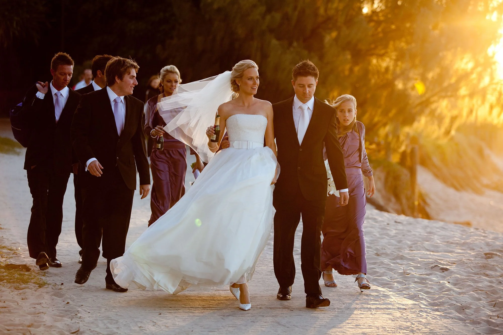 A bride in a wedding dress walking on a sandy beach at sunset, surrounded by bridesmaids and groomsmen, some holding champagne bottles, with trees and golden sunlight in the background.