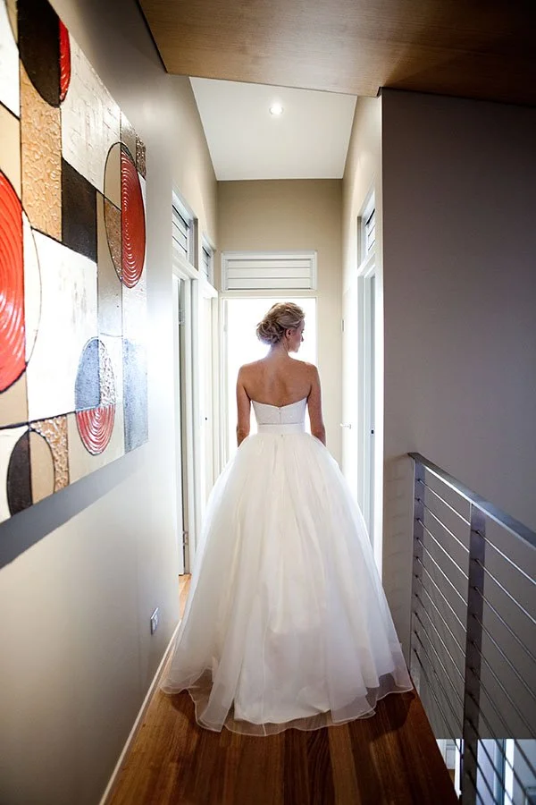 A woman in a white wedding dress standing in a hallway, looking toward a door with natural light from outside. Queensland Wedding 