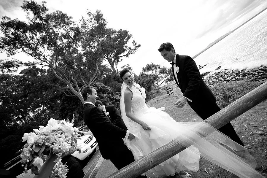 Black and white photo of a bride in a wedding dress and two grooms in tuxedos, smiling and laughing outdoors near water, with trees in the background and flowers on a vehicle in the foreground.