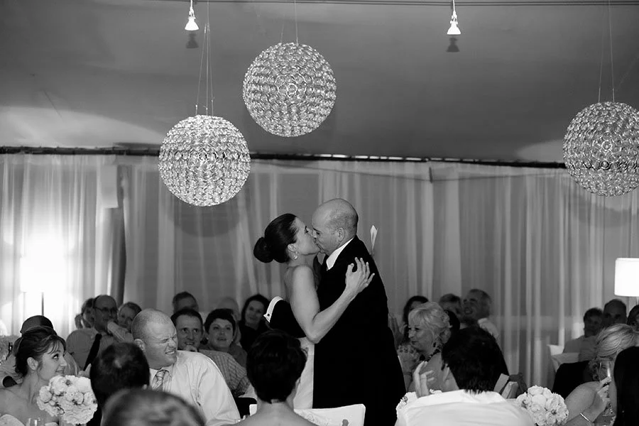 A bride and groom sharing a dance at their wedding reception, surrounded by seated guests, with decorative hanging lights in the background. Noosa Wedding 
