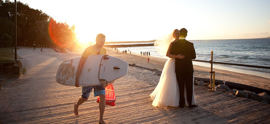 A bride and groom embrace on a beach at sunset, with a young man carrying a surfboard walking by in the foreground.