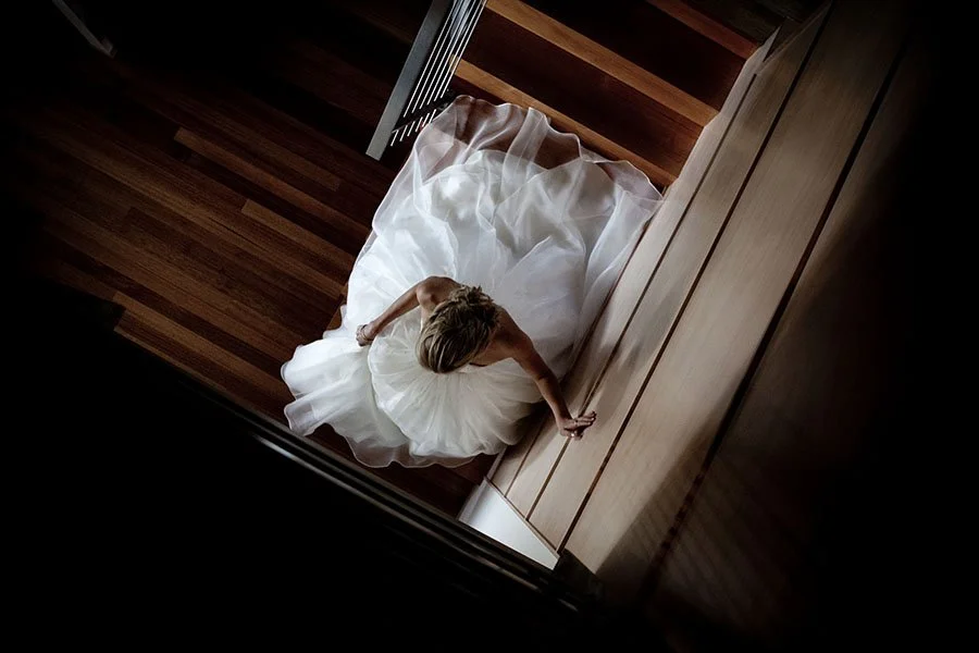 A woman wearing a white wedding dress sitting on a wooden floor near a staircase, viewed from above.