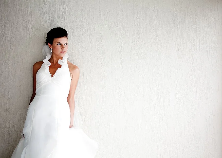 Bride in a white wedding dress with ruffled neckline, standing against a plain light-colored wall, looking to the side.