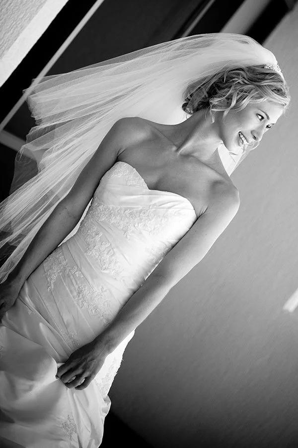 Black and white photo of a bride in a strapless wedding gown with lace details, smiling and looking to her right, wearing a veil and her hair styled in loose curls.