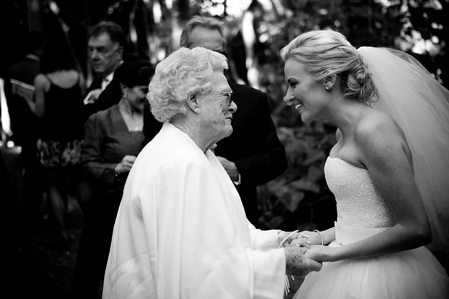 A bride and an elderly woman holding hands and smiling at each other, surrounded by other wedding guests in an outdoor setting.