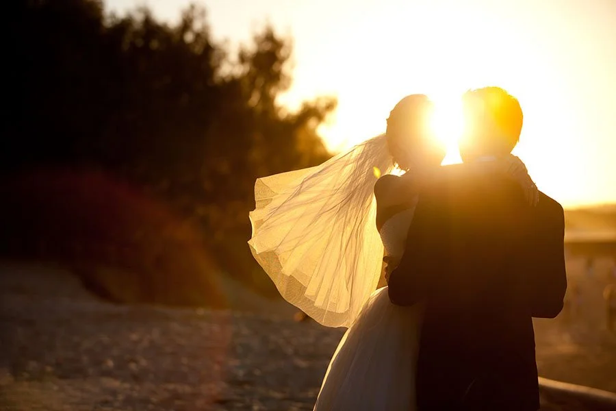 Silhouetted couple, a bride and groom, embracing at sunset on a beach, with the sun behind them casting a warm glow.