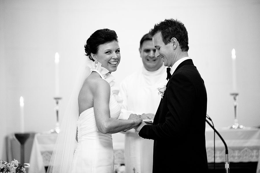 Black and white wedding photo of a bride and groom exchanging vows, with a minister witnessing in the background, candles on the altar.