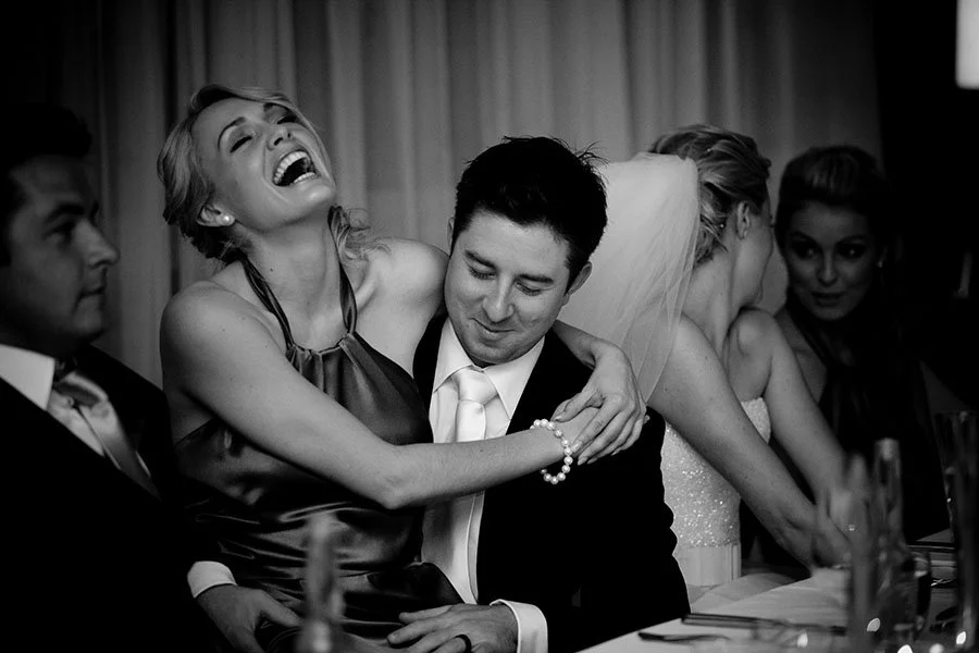 People at a wedding reception or celebration. A woman in a party dress is laughing and hugging a man in a suit and tie, who appears happy and slightly shy. Other guests are seated nearby, including a woman in a wedding dress and veil.