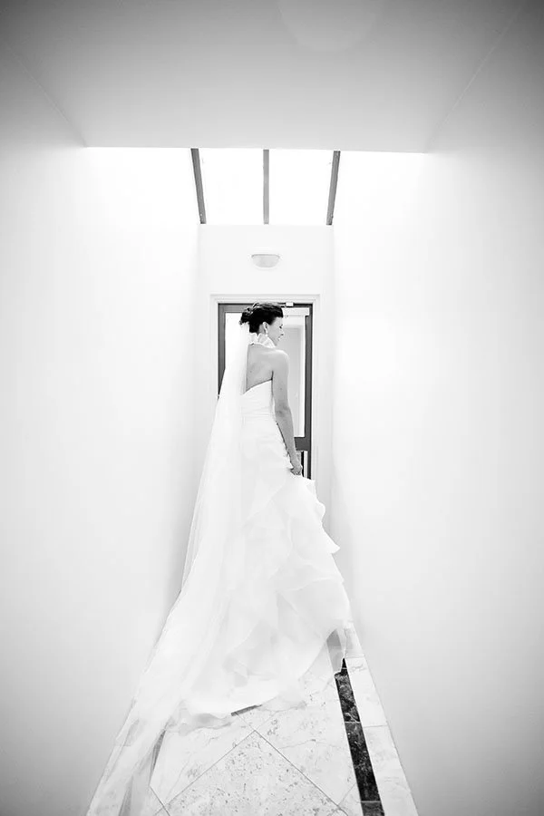 Bridal woman in wedding dress standing gracefully in a narrow hallway with bright walls and ceiling, holding her dress, facing away from the camera.