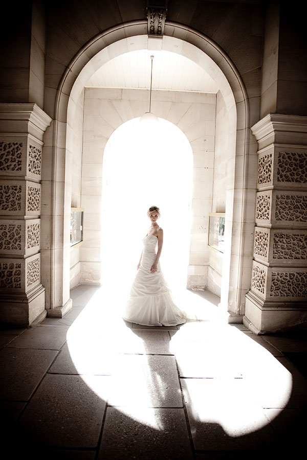 A woman in a wedding dress stands in an arched stone doorway, backlit by bright light, creating a silhouette effect.