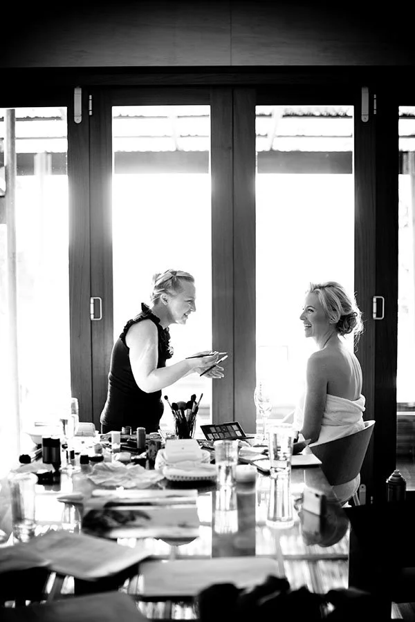 Two women in a bright room, one sitting and the other standing, smiling and talking, with makeup and beauty products on a table.