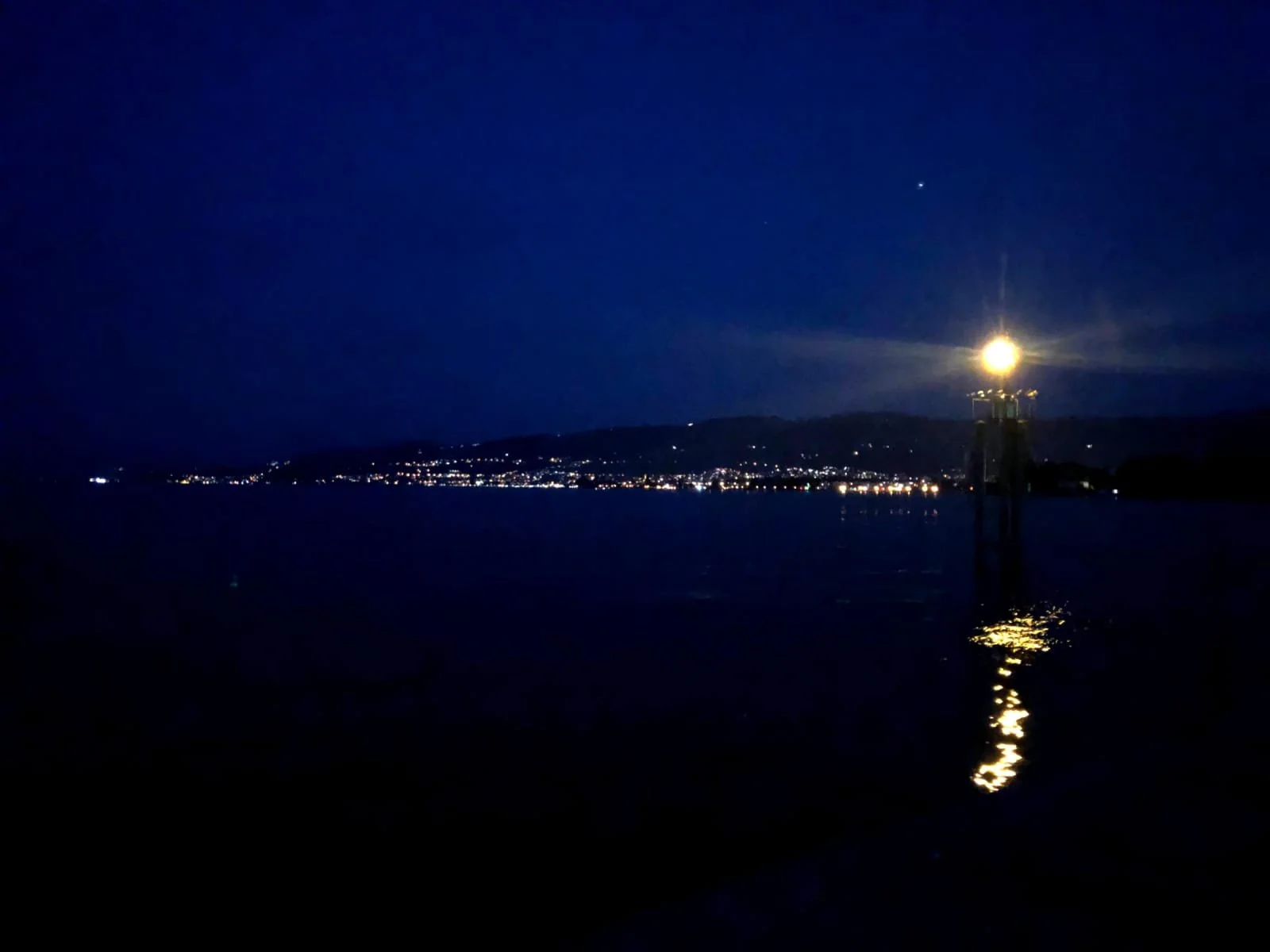 Nachtansicht eines Sees mit einem Leuchtturm, das Licht reflektiert auf dem Wasser, im Hintergrund beleuchtete Stadt am Ufer und dunkler Himmel mit wenigen Sternen, beruhigende Szene.
