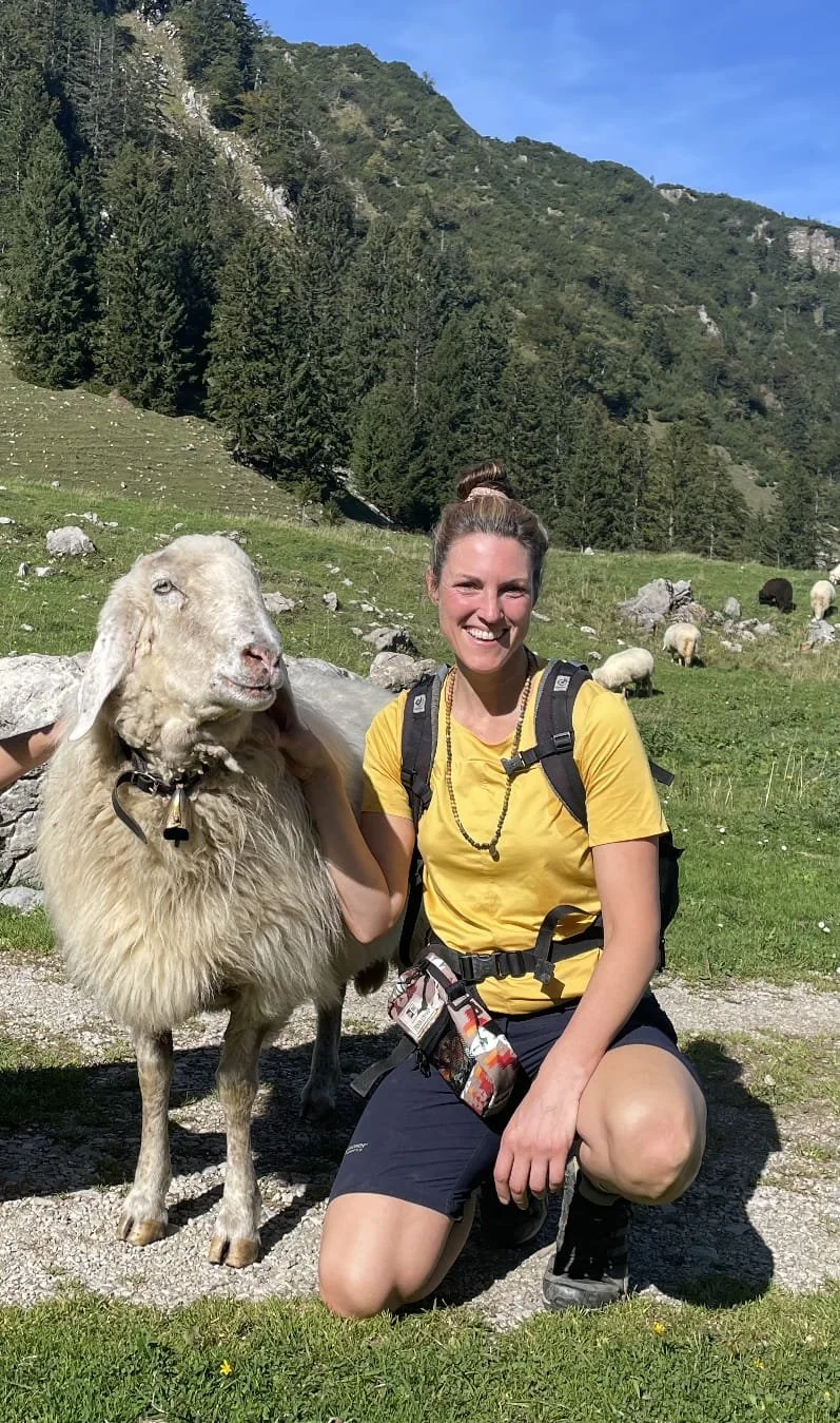 Verena in gelbem T-Shirt kniet neben einer Ziege auf einer Wiese in einer bergigen Landschaft mit Tannenbäumen im Hintergrund, sonniger Himmel.