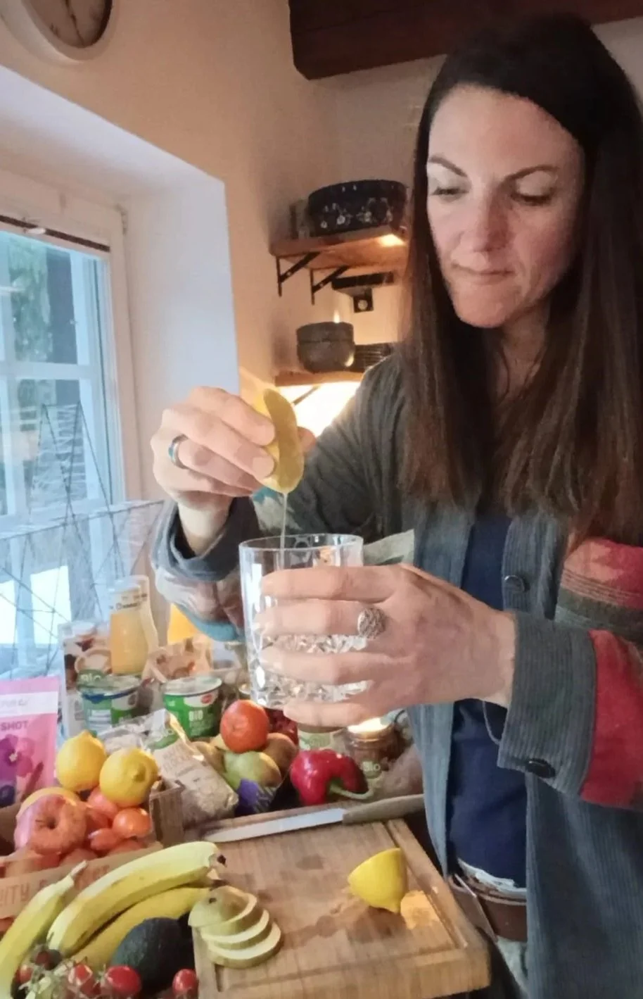 Verena presst eine Zitrone in ein Glas Wasser, umgeben von verschiedenen Obstsorten auf einer Küchenarbeitsplatte.