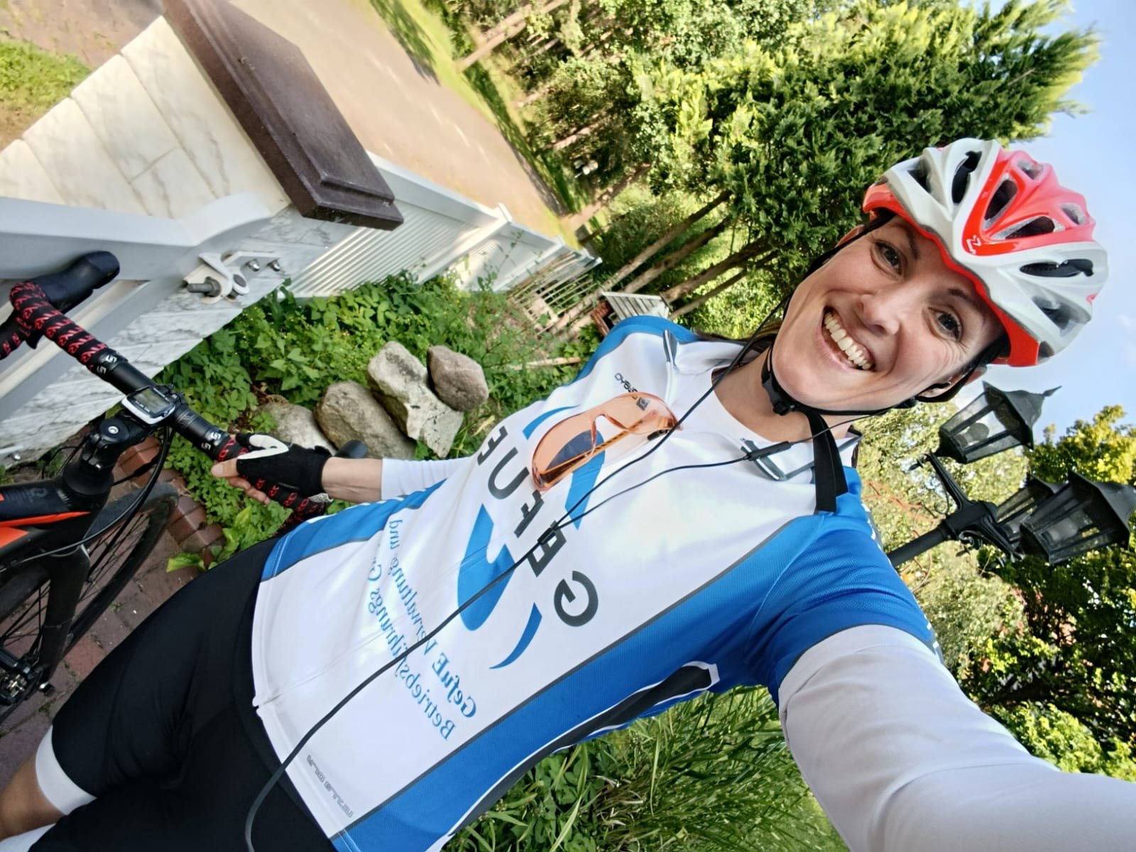 Jubelnder Radfahrer mit Helm und Radsportkleidung, Selfie im Freien, grüne Bäume und alte Laternen im Hintergrund.