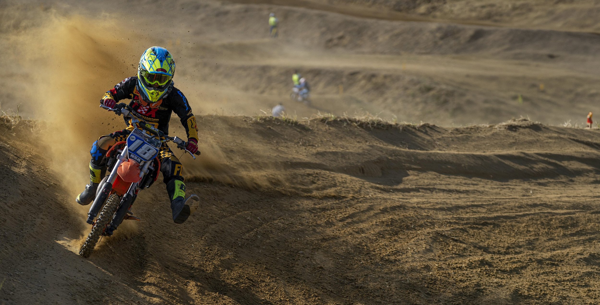Motorradfahrer in Schutzkleidung und Helm fährt auf einer Sandpiste mit Staub auf der Rennstrecke, andere Fahrer im Hintergrund.