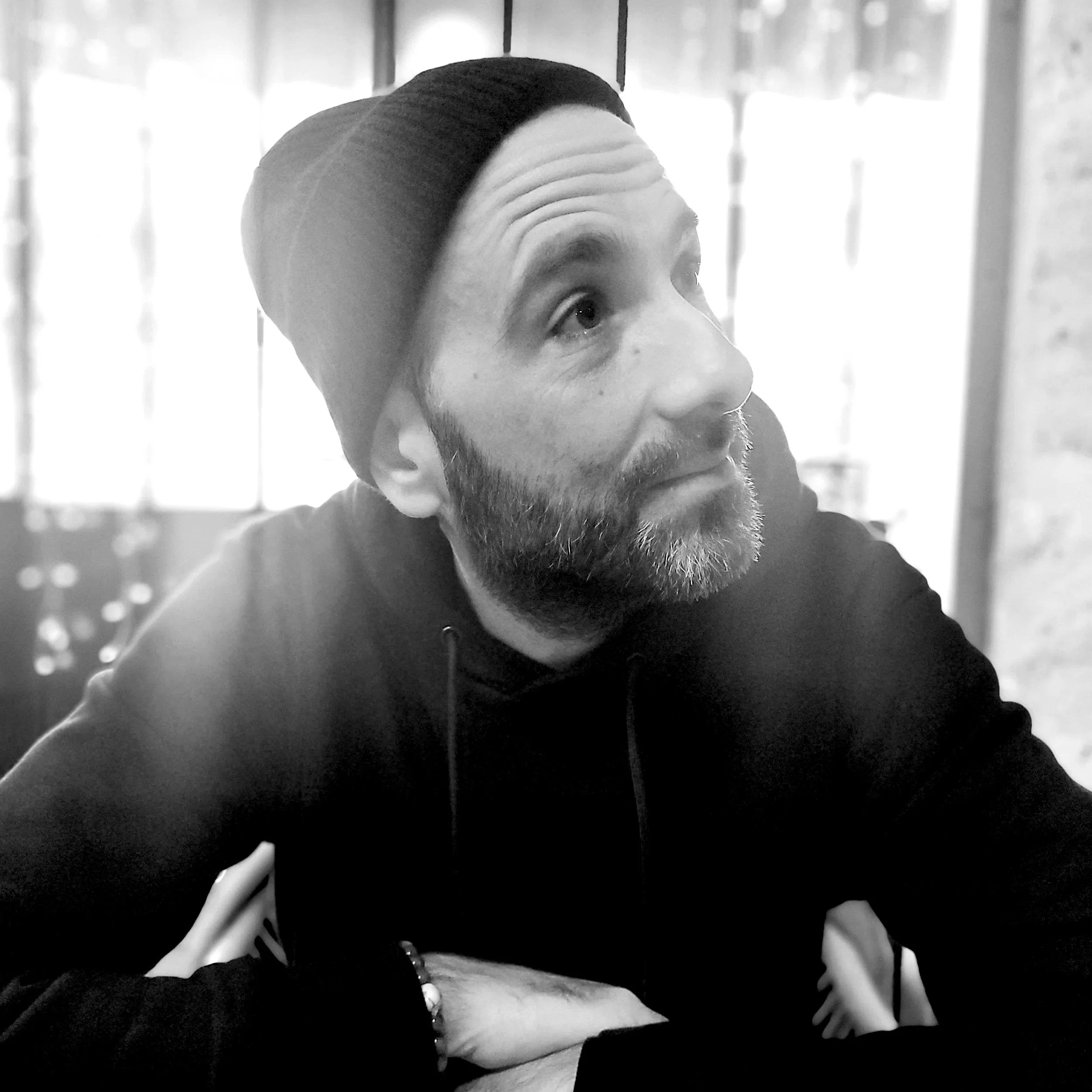 Un homme avec une barbe, portant un bonnet noir et un sweat noir, regarde vers le haut dans une pièce lumineuse avec des fenêtres en arrière-plan.