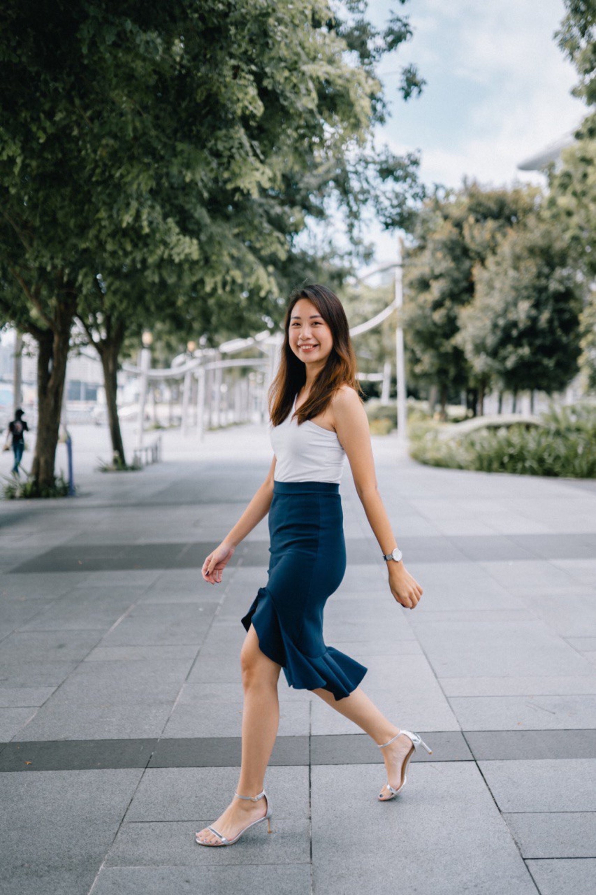 Agatha Neo, Propnex Singapore real estate agent, walking outdoors on a grey paved path with trees and a cityscape in the background. She is smiling and appears to be in motion, wearing high-heeled sandals.