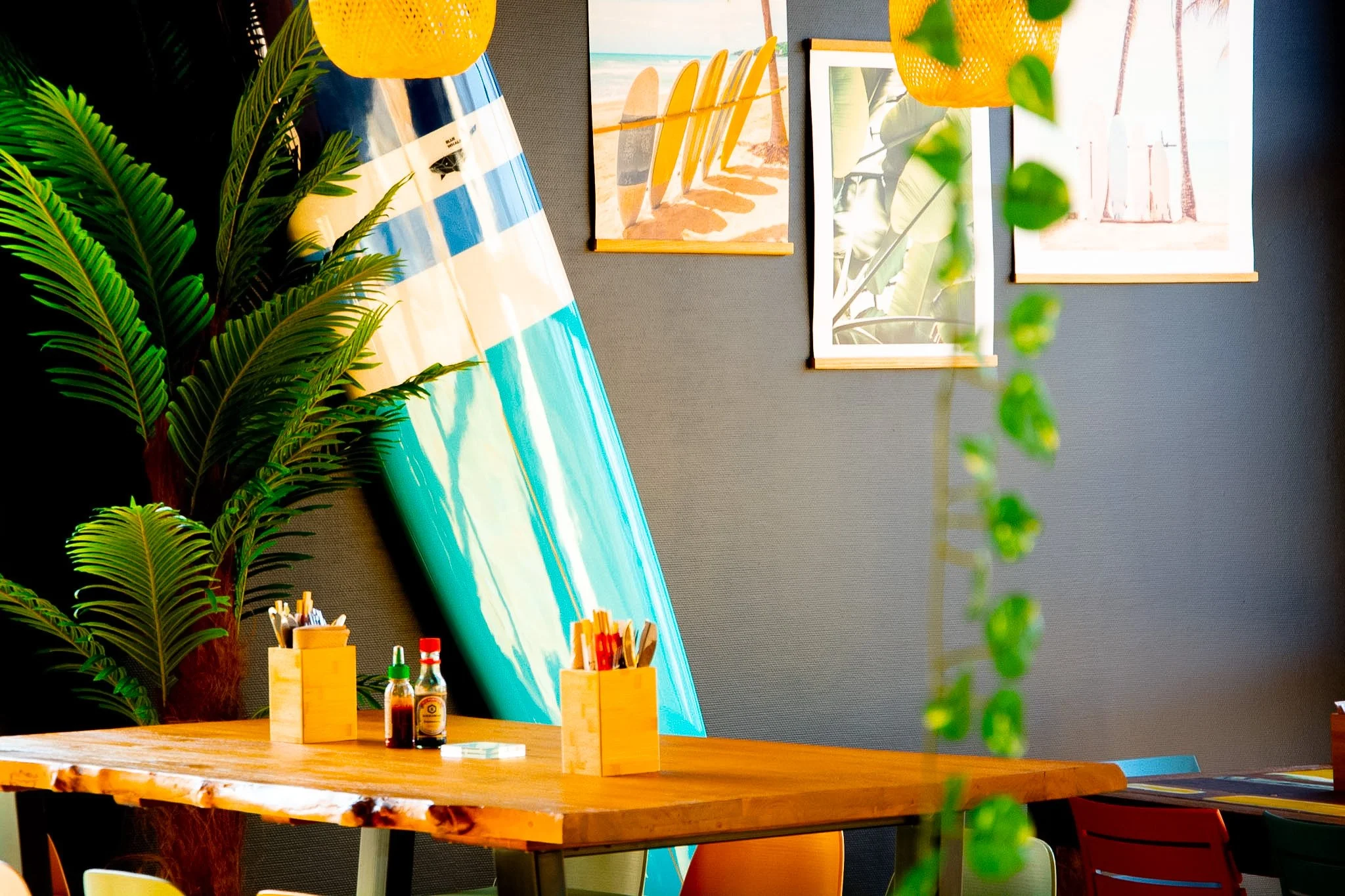A decorated indoor space with a large, colorful surfboard leaning against a dark wall, surrounded by green plants, with a wooden table set with condiment bottles and wooden containers, and framed beach-themed artwork hanging on the wall.