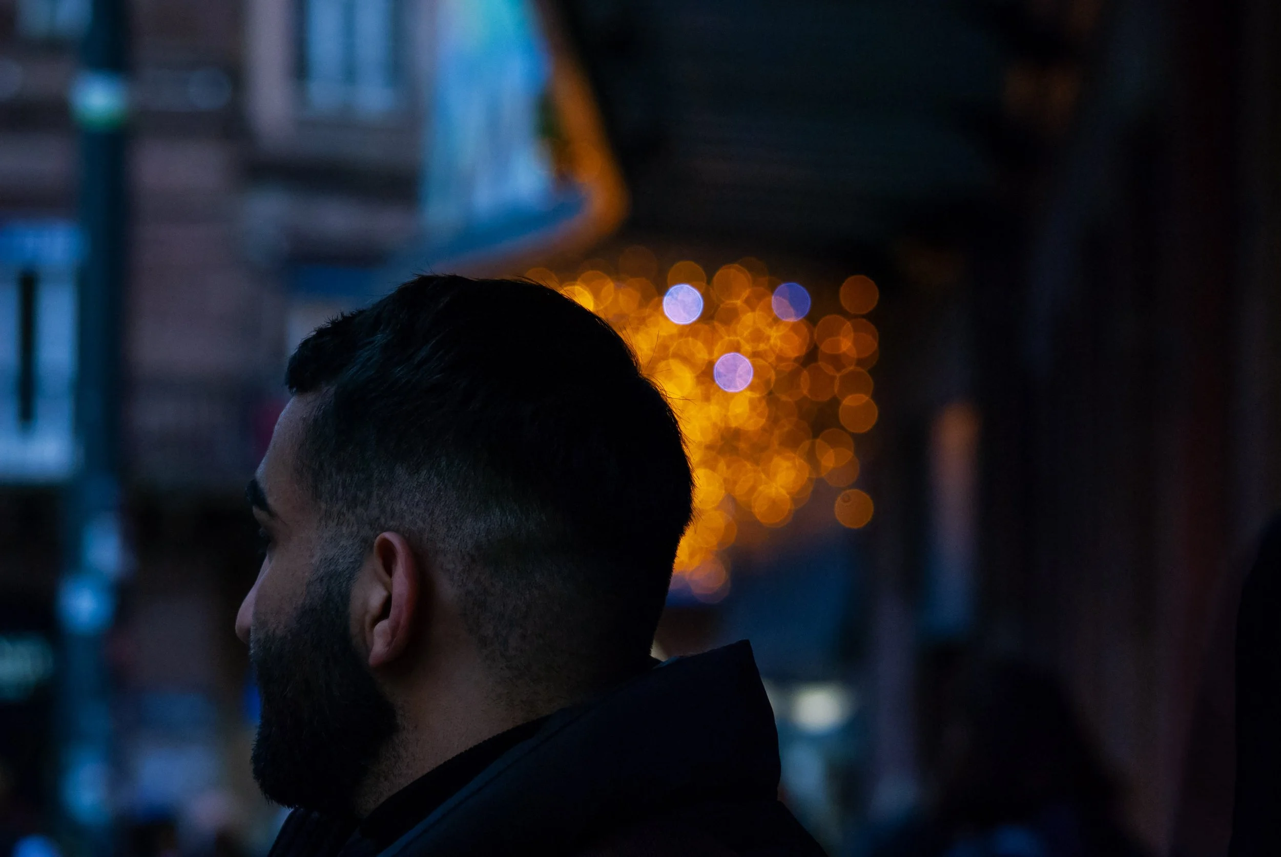 Side profile of a man with a beard and short dark hair, looking to the left, with warm orange and purple bokeh lights in the background during nighttime.
