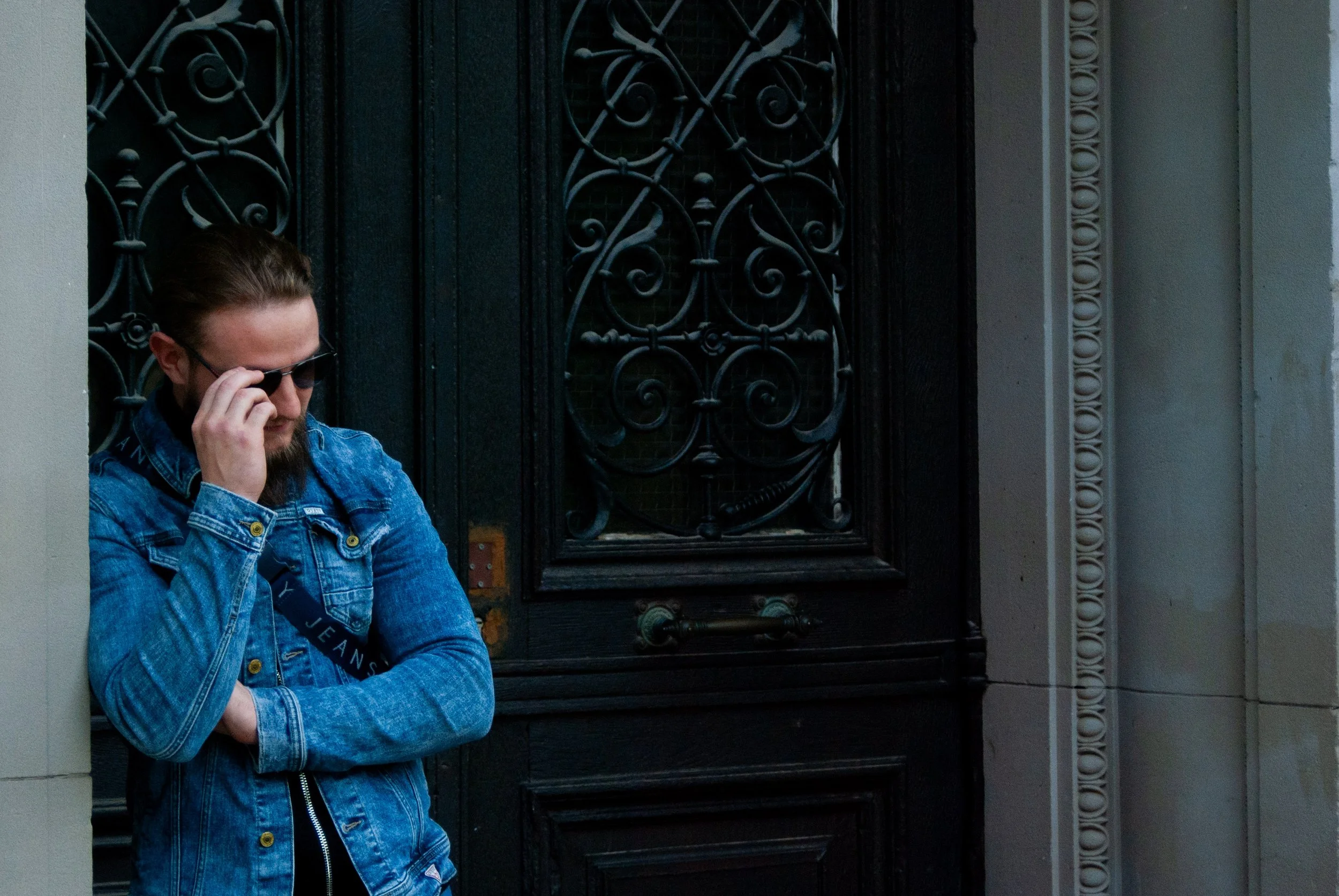 A man with long hair, sunglasses, and a beard, wearing a denim jacket, leaning against a beige wall and talking on a cell phone, standing in front of a decorative black gate with ornate metalwork.