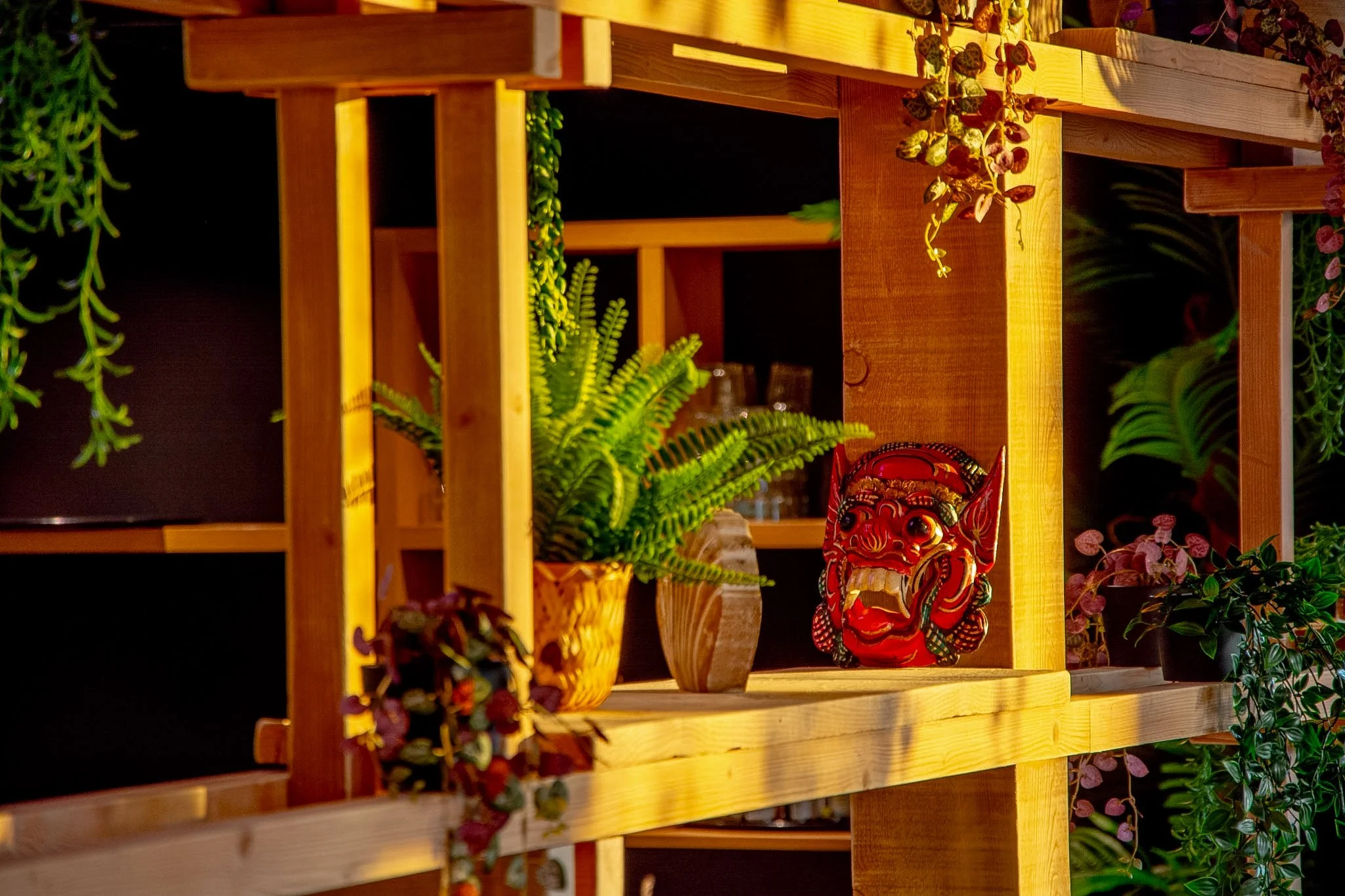 Wooden shelf with potted fern, small plants, and a red decorative mask with an angry face, surrounded by green plants and hanging vines.