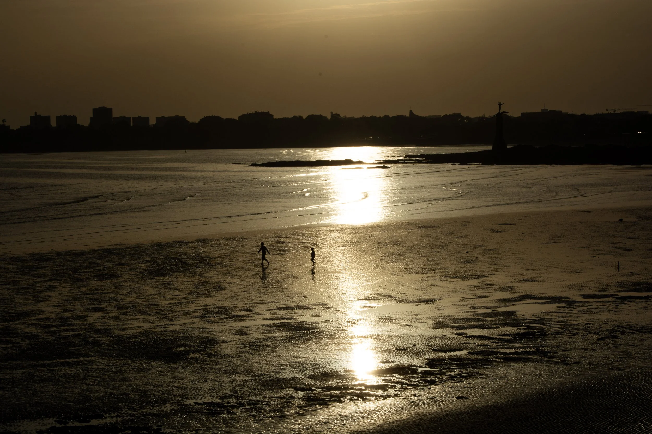 Beach at sunset with silhouetted children playing in the shallow water, city skyline in the background, reflected golden light on wet sand.