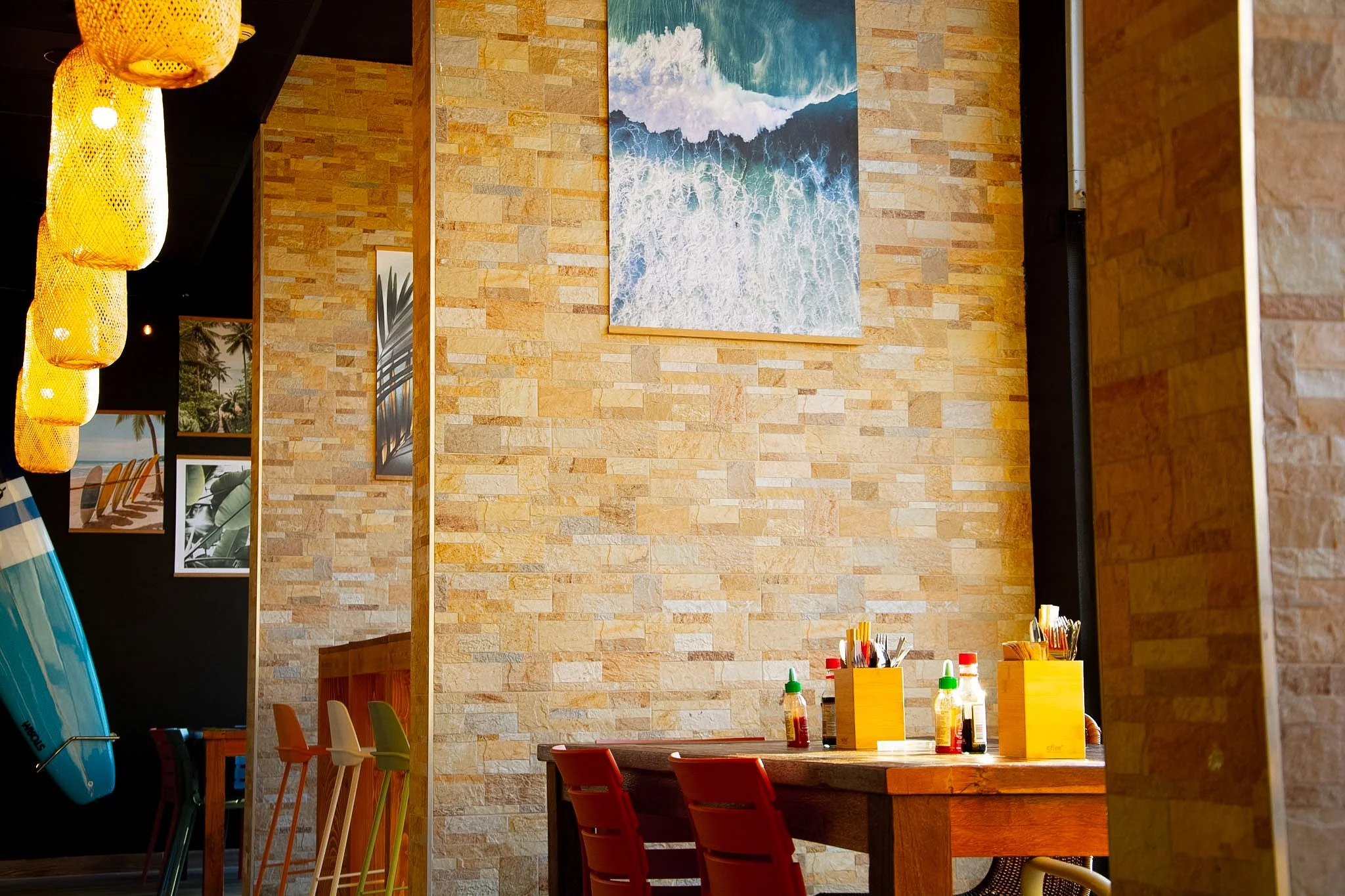 Interior of a restaurant with a tropical decor theme, featuring a wooden table with condiment bottles and utensils, colorful chairs, and wall paintings of beaches and ocean waves. Hanging lanterns on the left side and a surfboard mounted on the wall.