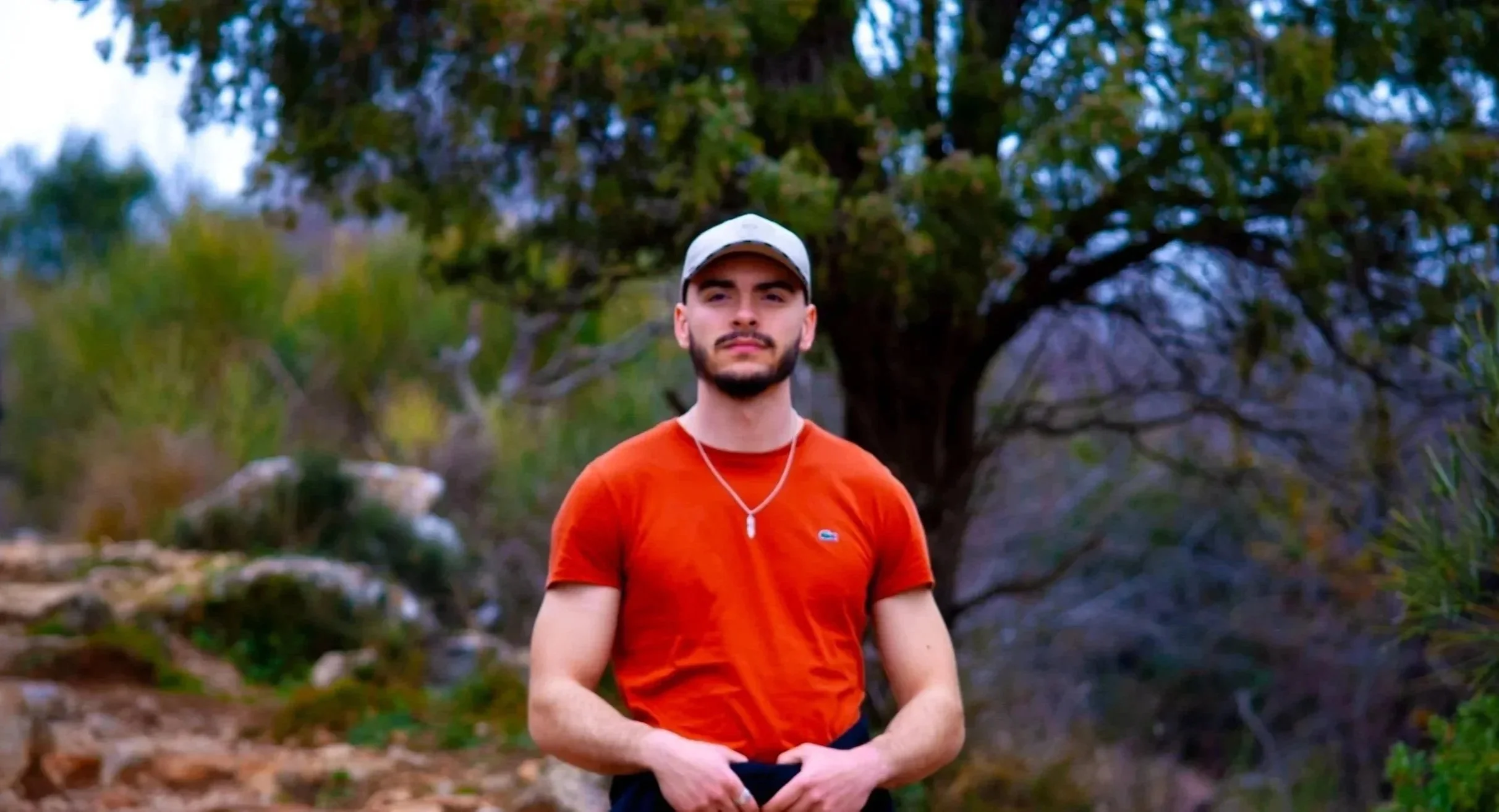 A young man with a beard, wearing an orange T-shirt, a white cap, and a necklace, standing outdoors in front of a large tree and greenery.
