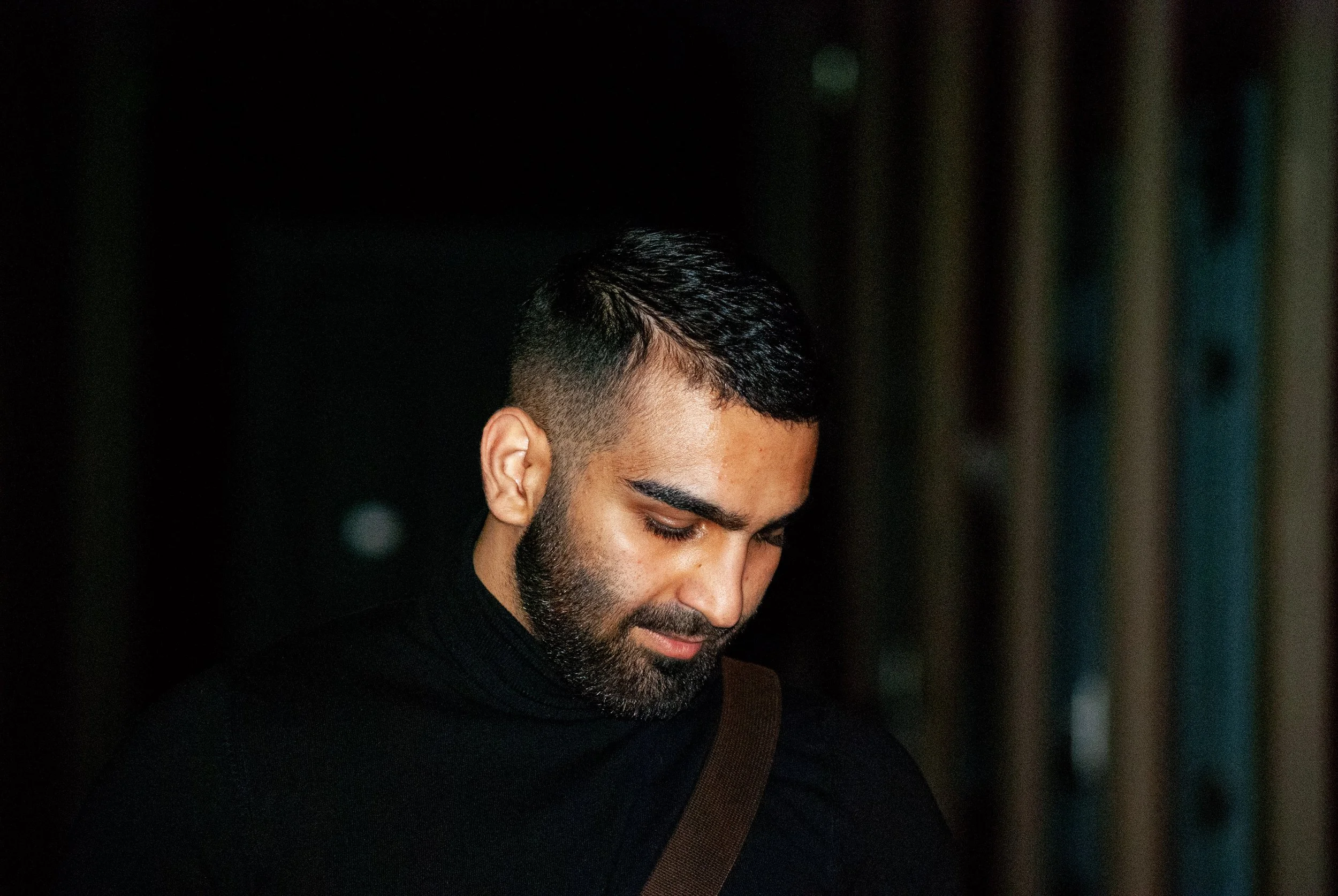 A man with short dark hair, a beard, and closed eyes looking downward, wearing a black shirt, in a dimly lit environment.