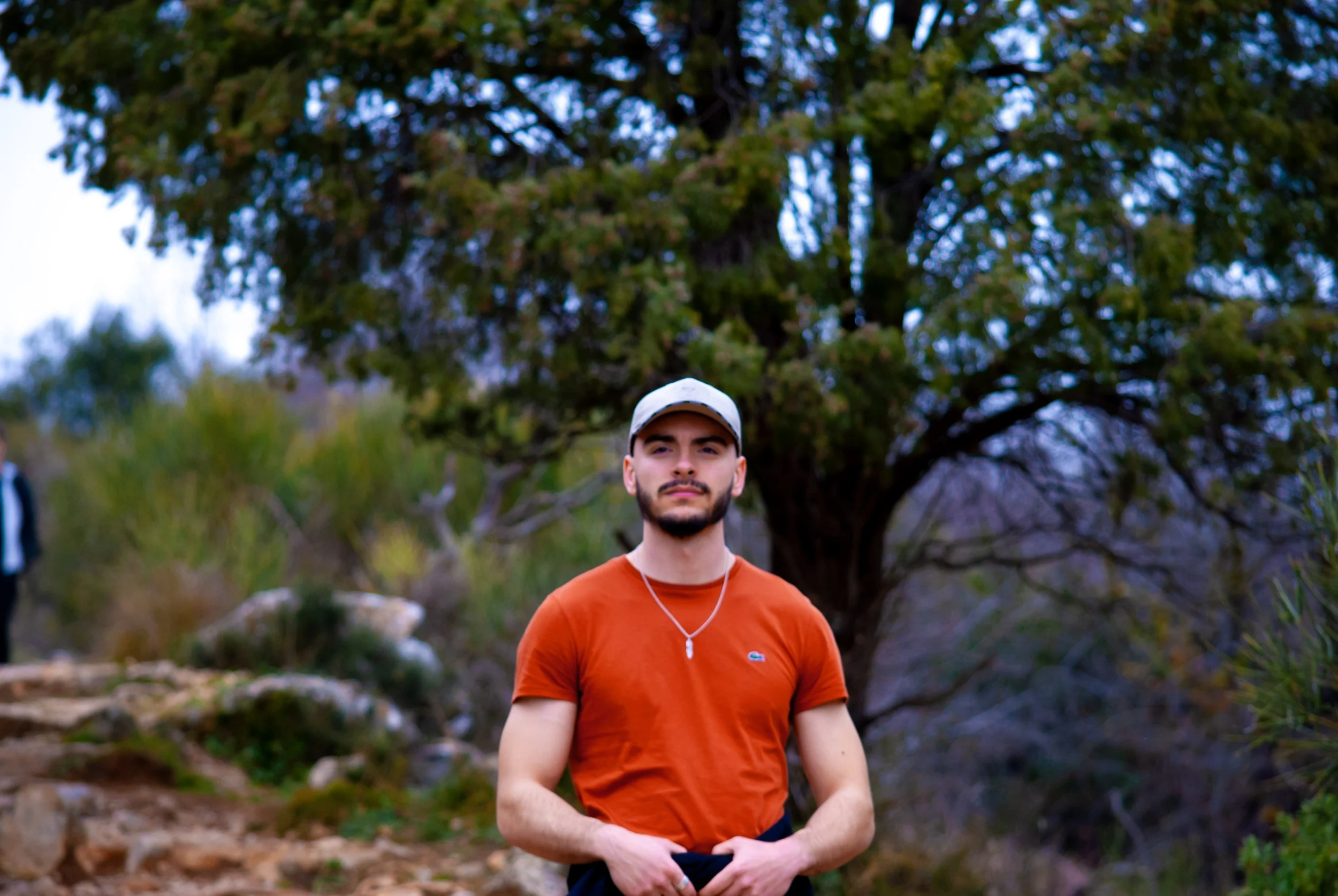 A young man with a beard, wearing a light gray cap, an orange t-shirt, and a necklace, standing outdoors in a natural setting with trees and rocks in the background.