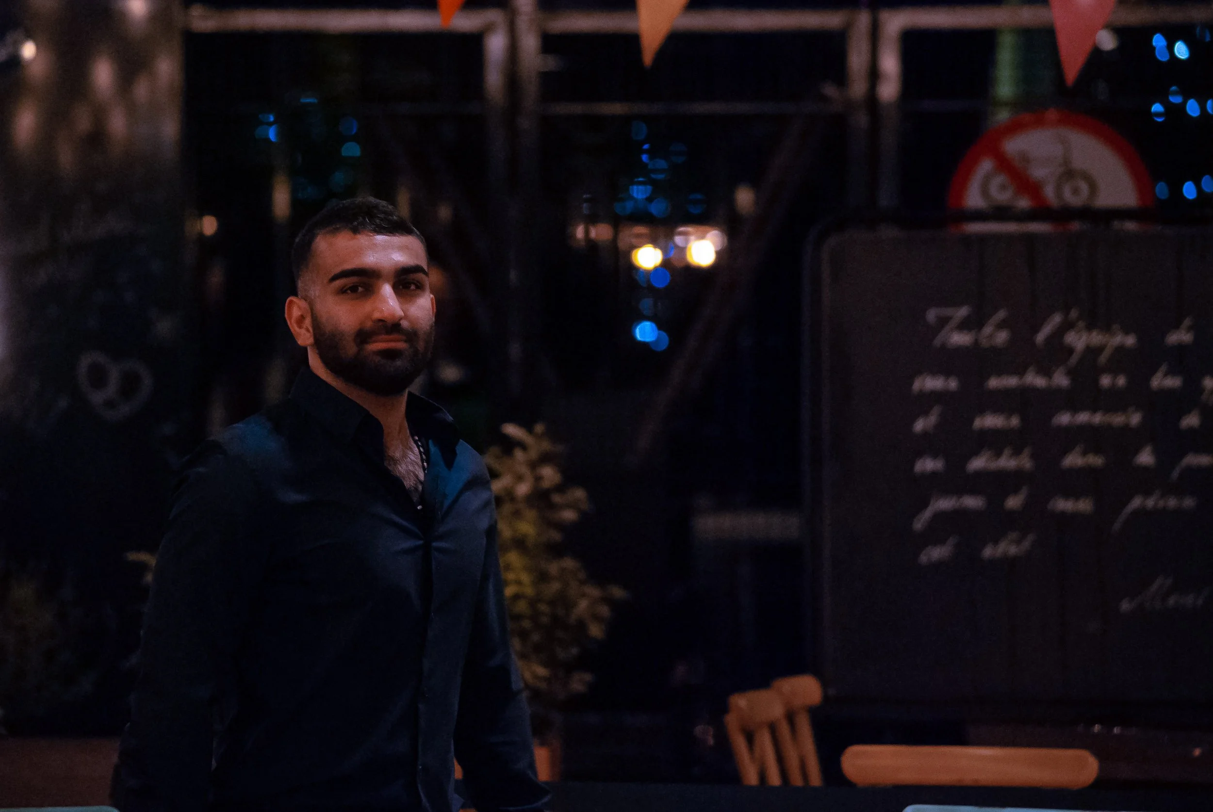 A man with a beard wearing a dark shirt standing in a dimly lit outdoor setting at night, with blurred lights and a chalkboard menu in the background.