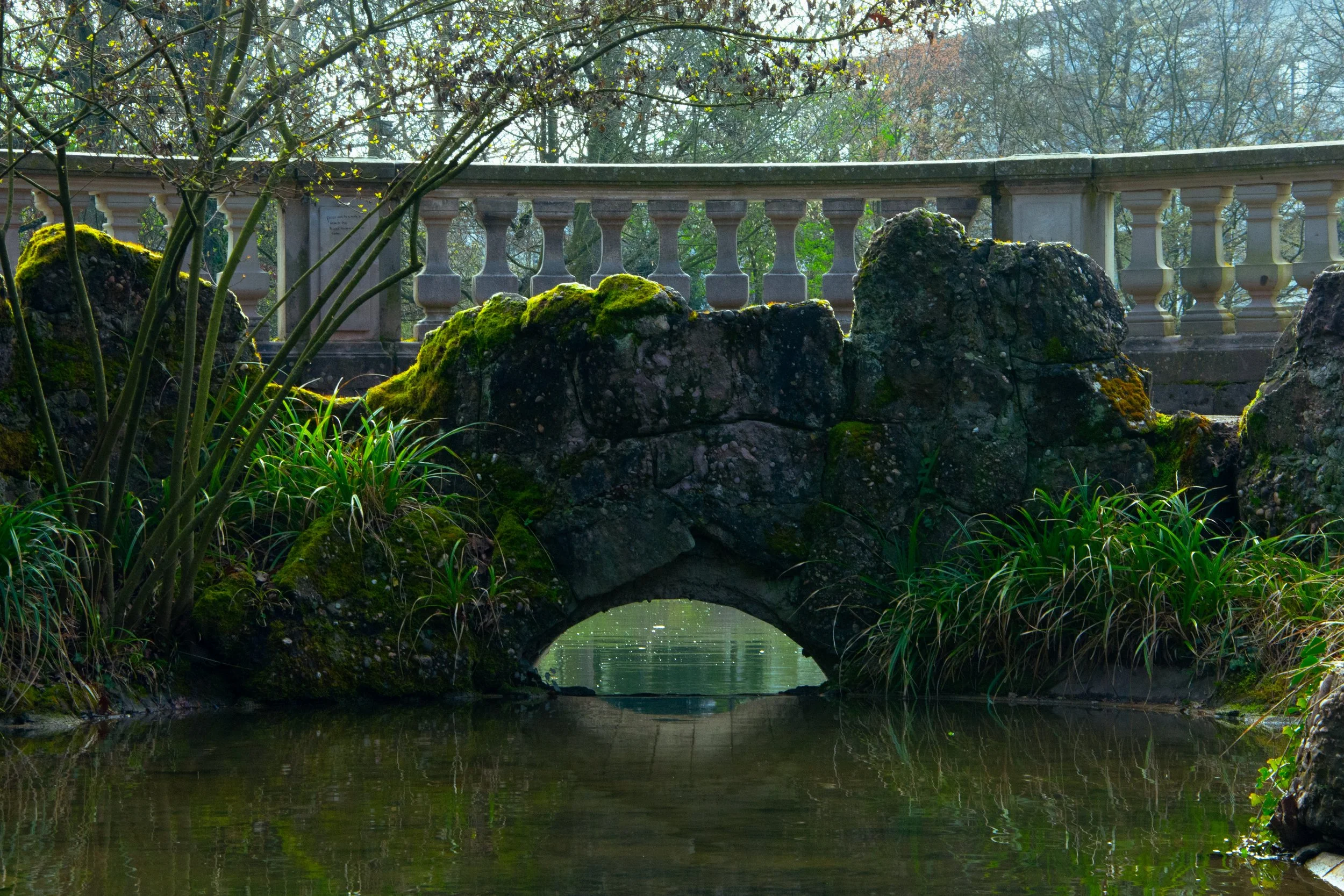 A small stone bridge with mossy rocks over a calm body of water, surrounded by green plants and trees, with a stone railing in the background.
