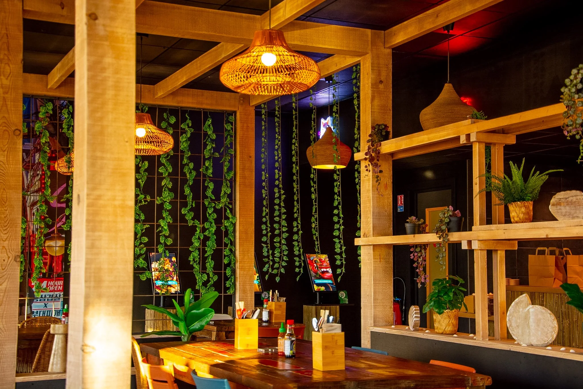Interior of a restaurant or cafe with wooden beams, hanging wicker lamps, decorative hanging plants, and a wooden shelf with potted plants and decorative items.