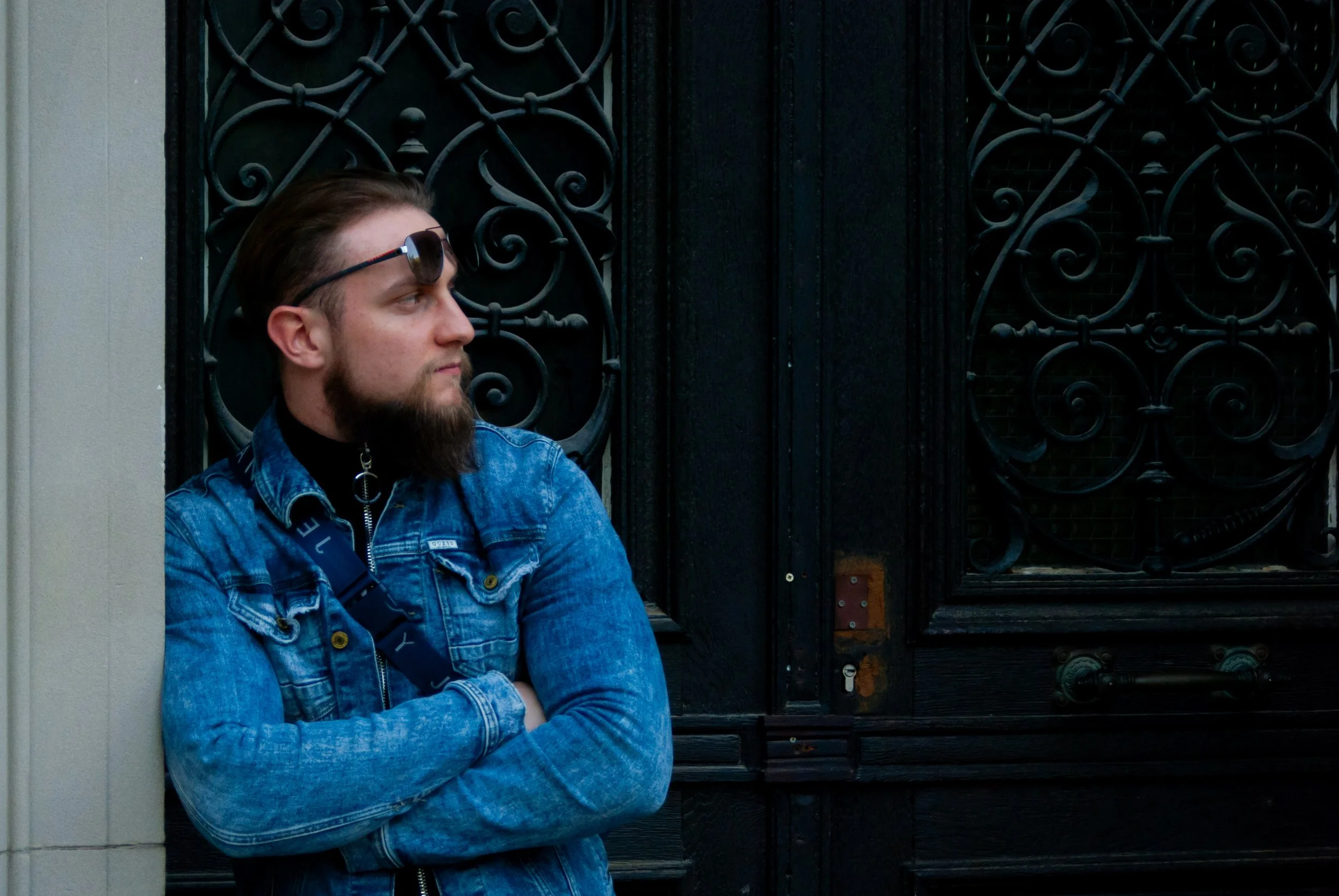 A man with a beard and sunglasses on his head leaning against a wall with arms crossed, standing near an iron gate.
